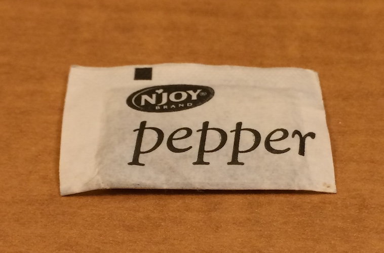 A single individual paper packet of pepper, no longer contained within a pepper shaker.